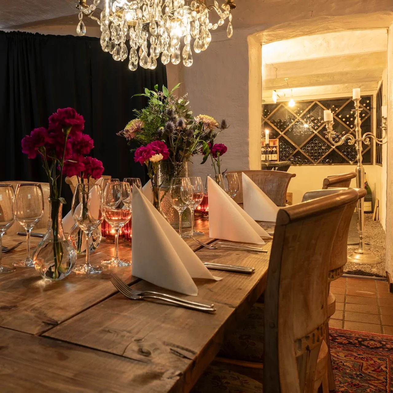 A wooden dining table, elegantly set with wine glasses, folded napkins, and flower vases, sits under a crystal chandelier; background features a warmly lit wine rack.