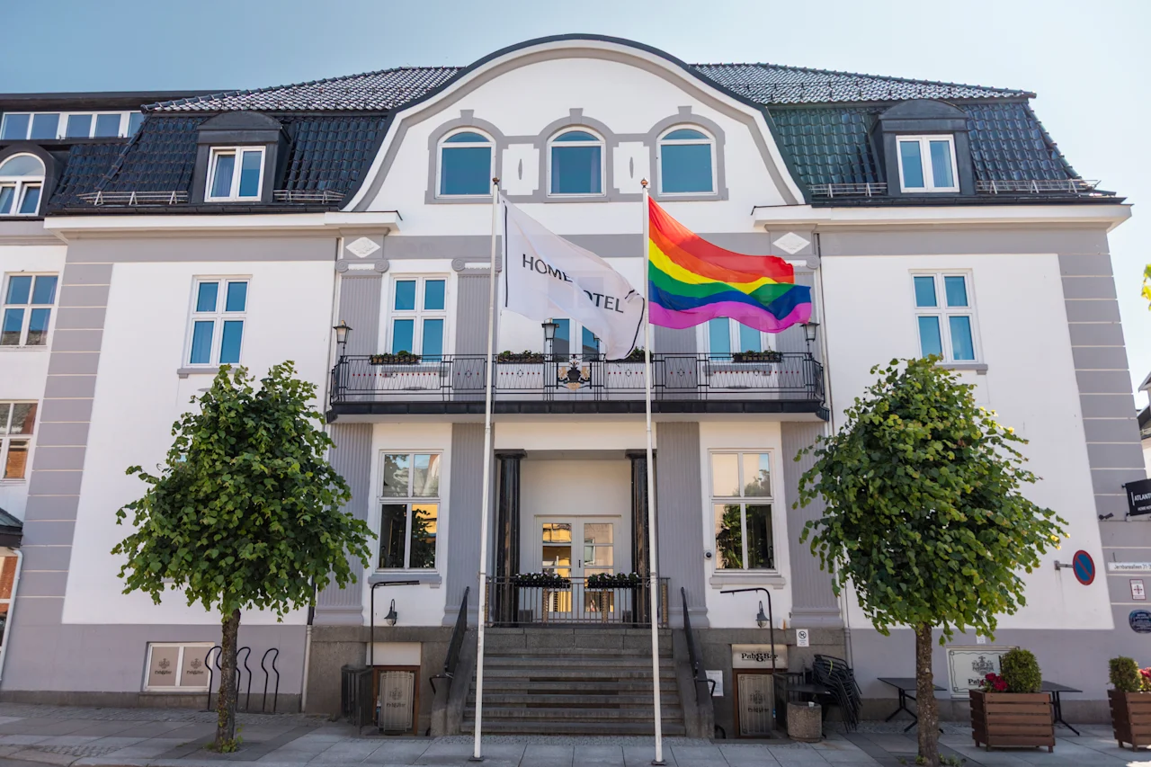 En indbydende hvid bygning med mørkt tag, der viser et hvidt flag med HOME TEL og et levende regnbueflag.