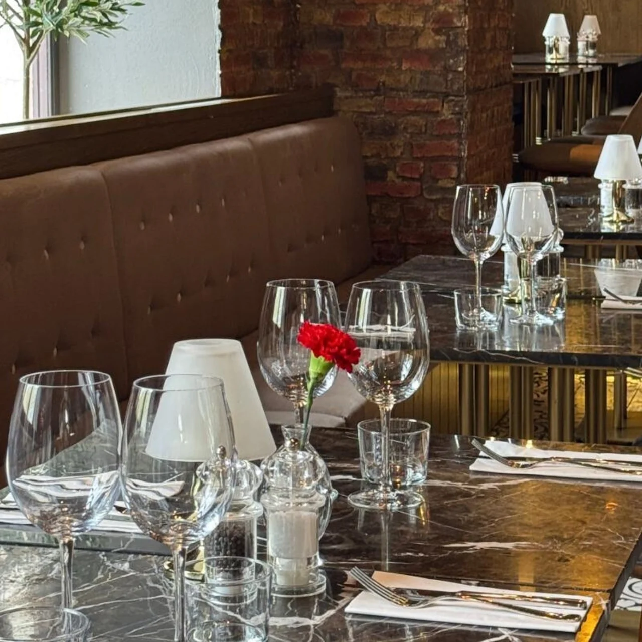 Elegant spisebord, dekoreret med tomme vinglas og en rød blomst, placeret i en restaurant med brune lædersæder og murstensvægge.