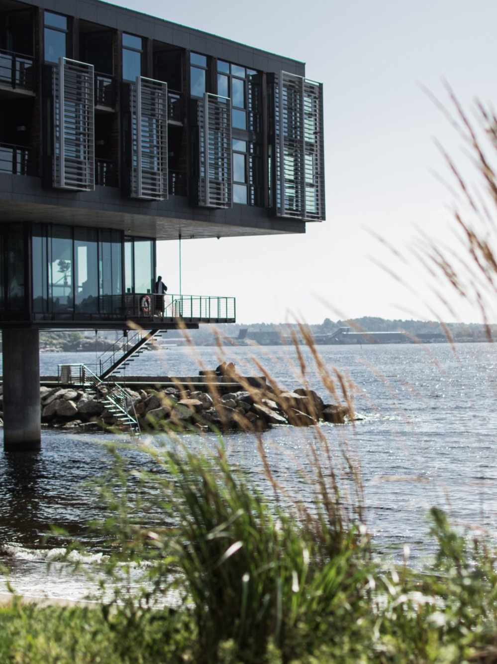 A modern building on stilts over water, with a person on the balcony and another by the shore, against a backdrop of industrial structures.