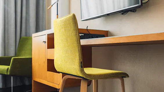 A green chair sits by a wooden desk with a black lamp. A wall-mounted TV is above the desk. The room has gray curtains and a textured rug.