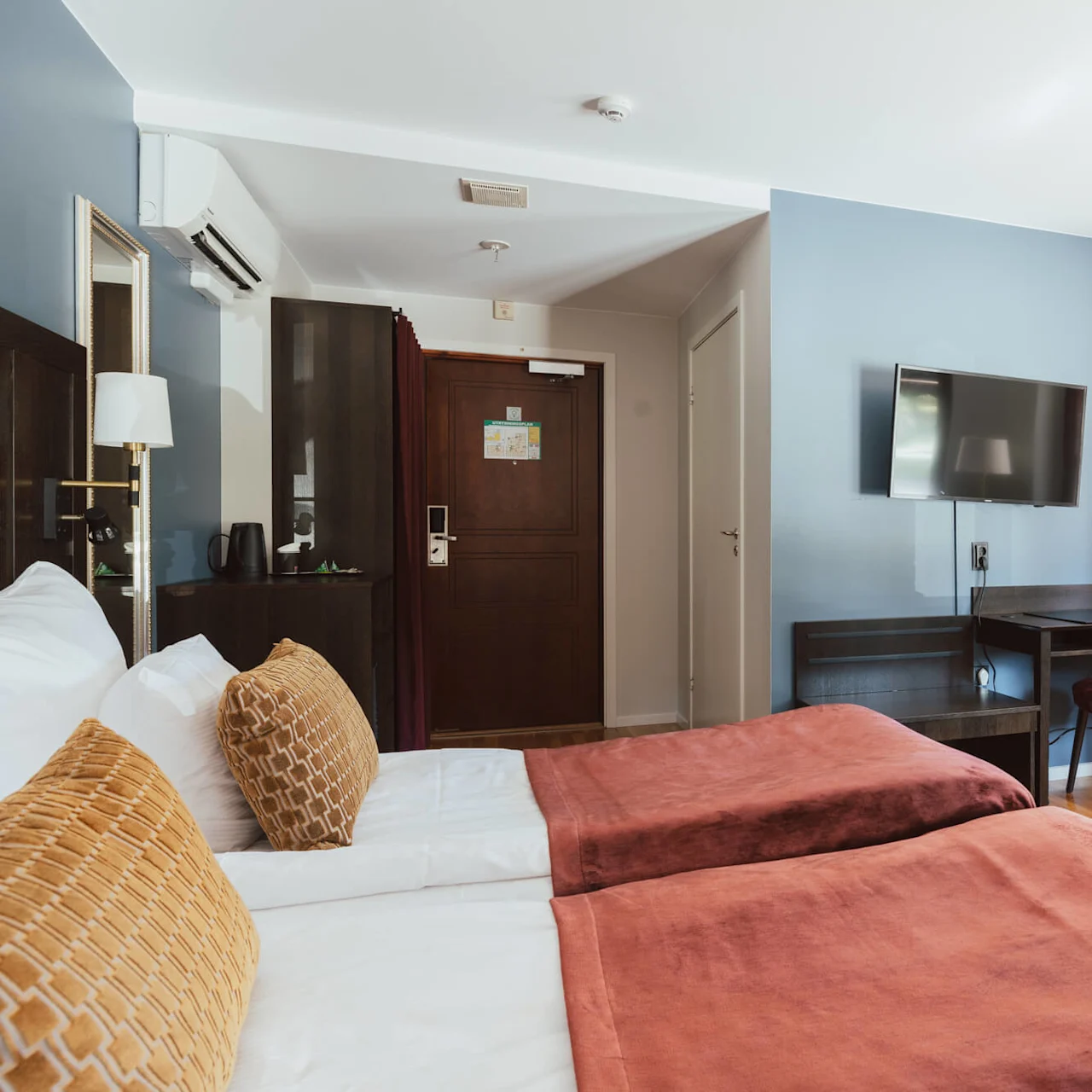 Et ryddig hotellrom med to senger, skrivebord og TV, opplyst av naturlig lys fra et vindu.