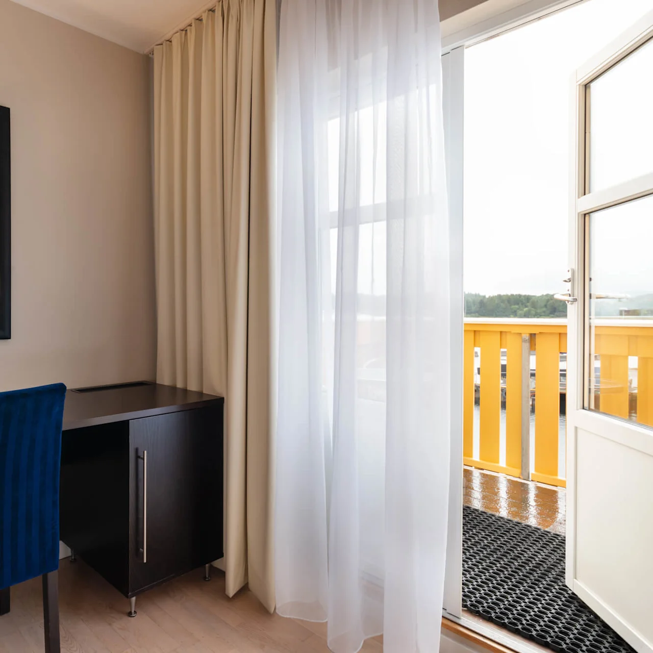 A black desk with a blue chair stands in a room beside a lamp and mirror, next to sheer curtains revealing an open door leading to a balcony with yellow railing.