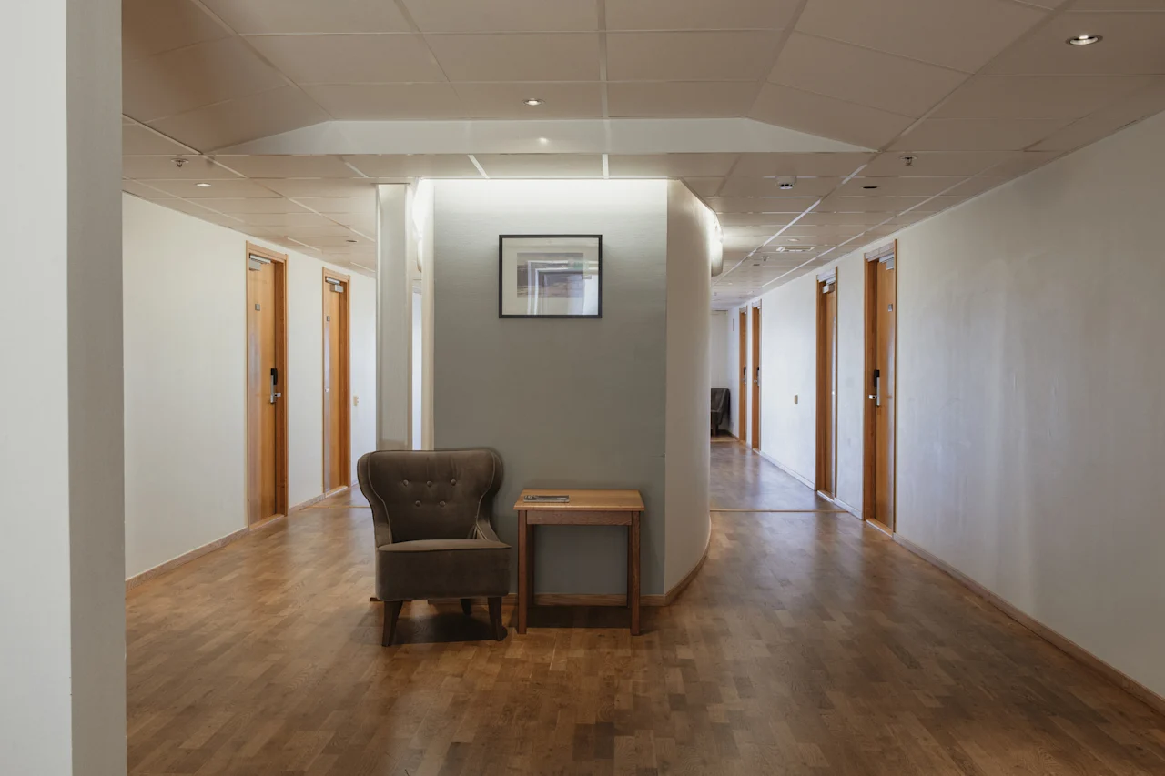 Hotellikäytävä, jossa on puulattiat, useita ovia, viihtyisä nojatuoli, pieni pöytä ja kehystetty kuva seinällä.