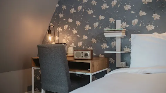 A cozy bedroom features a wooden desk with a lit lamp, radio, and books against a floral patterned wall, accompanied by a grey chair and nearby bed with white bedding.