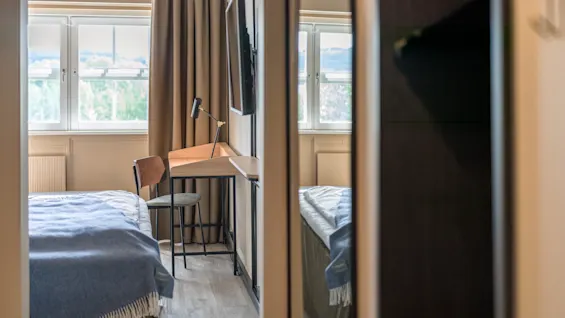 A wooden desk with a lamp and chair stands near a window with curtains. A bed with a blue blanket is in the foreground, reflecting in a mirror.