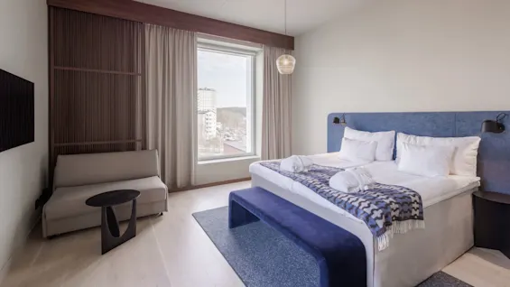 A modern bedroom with a neatly made bed, a bench, sofa, and window with a view of buildings.