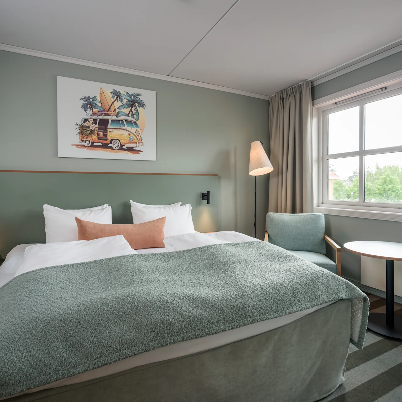 A neatly made bed with green covers is centered in a modern hotel room, flanked by small nightstands. A floor lamp stands nearby, and a window shows greenery outside.