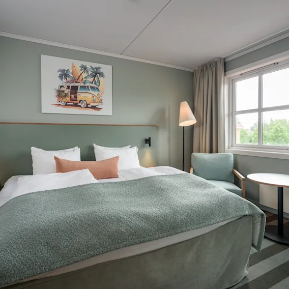 En pent senger med grønt sengeteppe står i sentrum av et moderne hotellrom, flankert av små nattbord. En gulvlampe står i nærheten, og et vindu viser grønt utenfor.