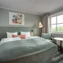 En pent senger med grønt sengeteppe står i sentrum av et moderne hotellrom, flankert av små nattbord. En gulvlampe står i nærheten, og et vindu viser grønt utenfor.