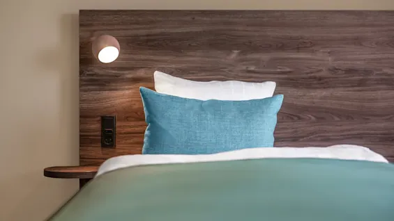 A bed with blue and white pillows rests against a wooden headboard. A small reading lamp is illuminated on the left, creating a cozy atmosphere.
