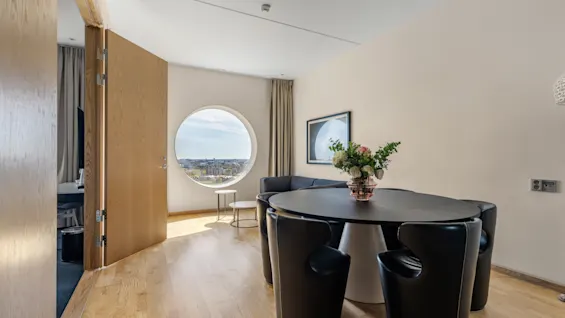 Et moderne hotelværelse med et rundt vindue, der tilbyder byudsigt, et spisebord med stole og et behageligt siddeområde.