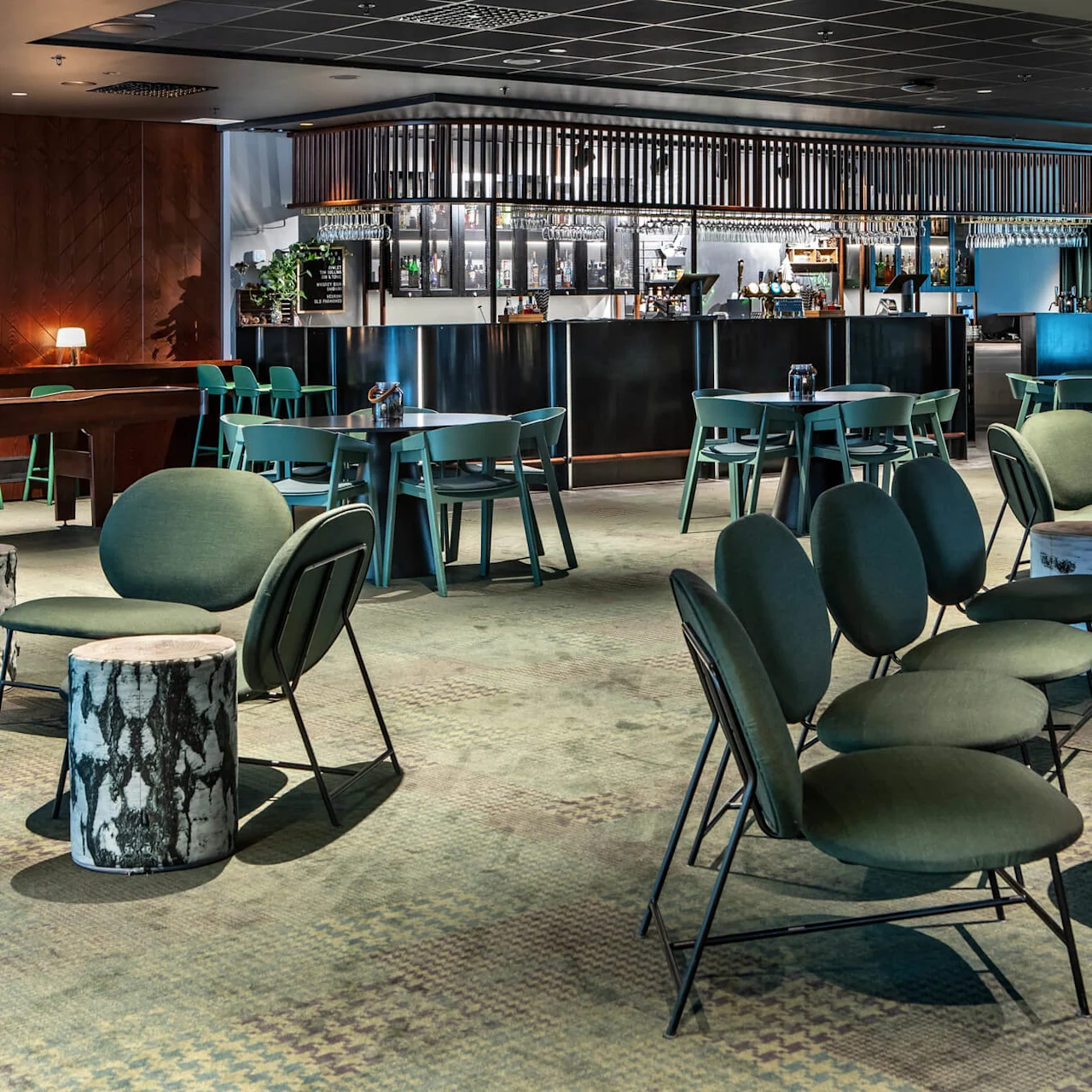 A modern lounge with green chairs surrounds a central bar. Round tables and tree-stump stools populate the carpeted area, featuring an illuminated drink display behind the bar.