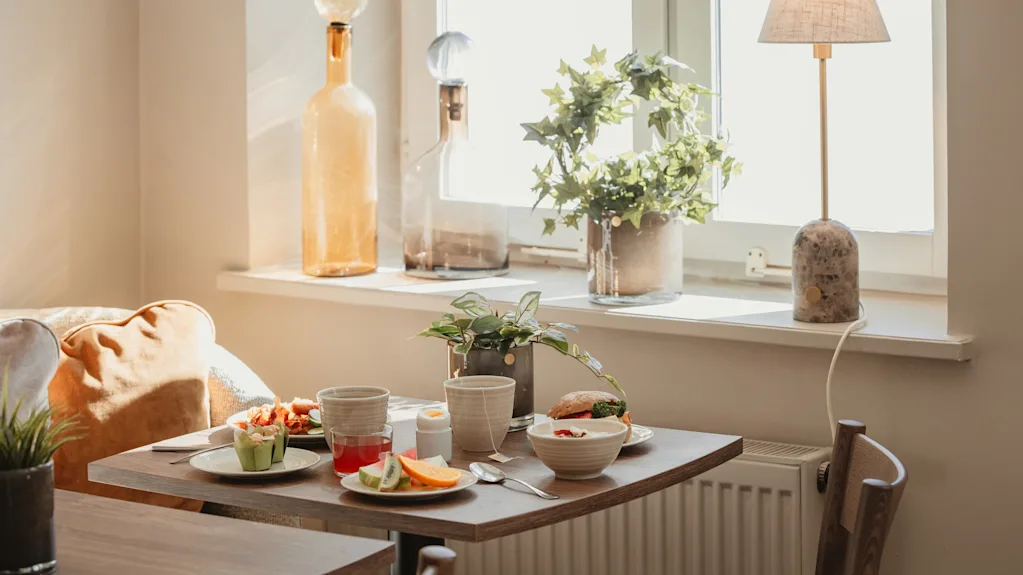 A cozy breakfast spread on a table by a sunlit window, with fresh fruit, a sandwich, and drinks.