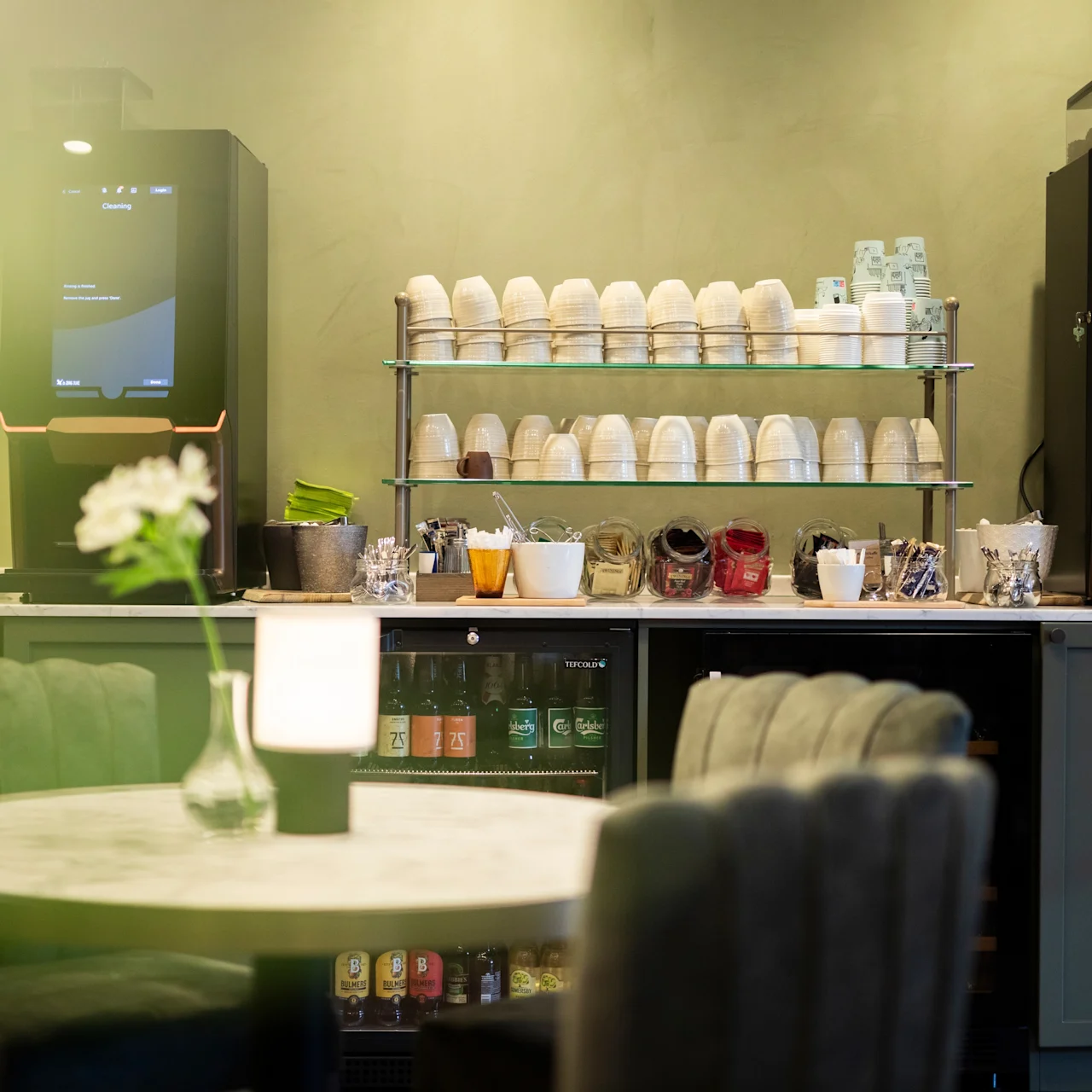 Coffee machines stand on a counter. Shelves hold stacked cups, with various condiments below. A round table with soft chairs and a flower vase is in the foreground.