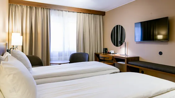 To pænt opredte enkeltsenge står side om side i et hotelværelse med beige vægge; et skrivebord og stol er placeret under et vægmonteret TV.
