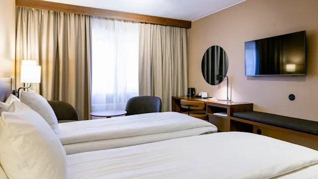 Två prydligt bäddade enkelsängar står sida vid sida i ett hotellrum med beige väggar; ett skrivbord och stol är placerade under en väggmonterad TV.