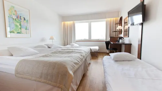A hotel room with two large beds and a small bed. A desk with a chair and lamp sits beneath a wall-mounted TV. A colorful painting hangs on a white wall.