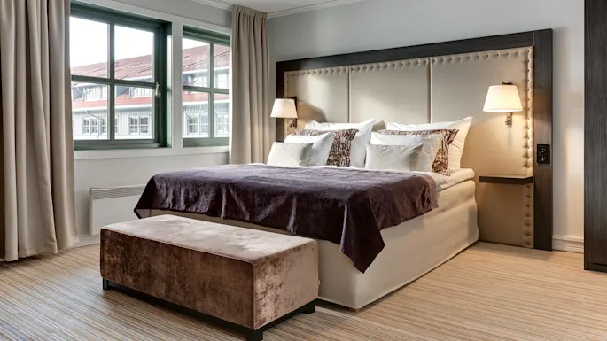 A bed with multiple pillows and a dark blanket stands against a cushioned headboard. Two lamps adorn the sides. Windows with beige curtains overlook neighboring rooftops.