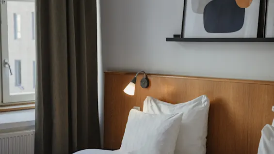 Viihtyisä hotellihuone Home Hotel Uppsalassa, jossa on sänky, valkoiset liinavaatteet, puinen sängynpääty, seinävalaisin ja abstraktia taidetta.
