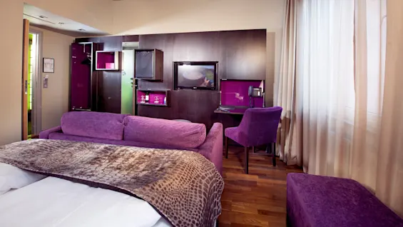 Lilla møbler i et koselig hotellrom med seng, sofa og skrivebord, parkettgulv og lyse gardiner.