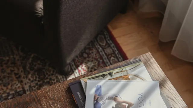 Böcker ligger staplade på ett träbord, en med titeln "Yoga" visar en kvinna i yogaposition. En mörk fåtölj och mönstrad matta antyder ett mysigt vardagsrum.
