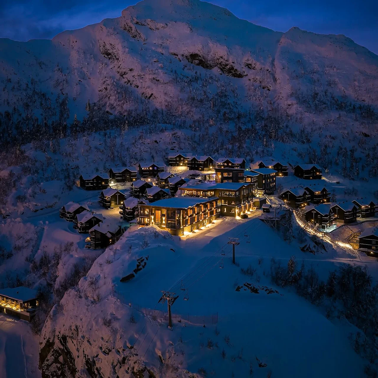 Chalet-style buildings glow with warm lights, nestled in a snowy mountainous landscape during twilight, surrounded by snow-covered peaks and trees.