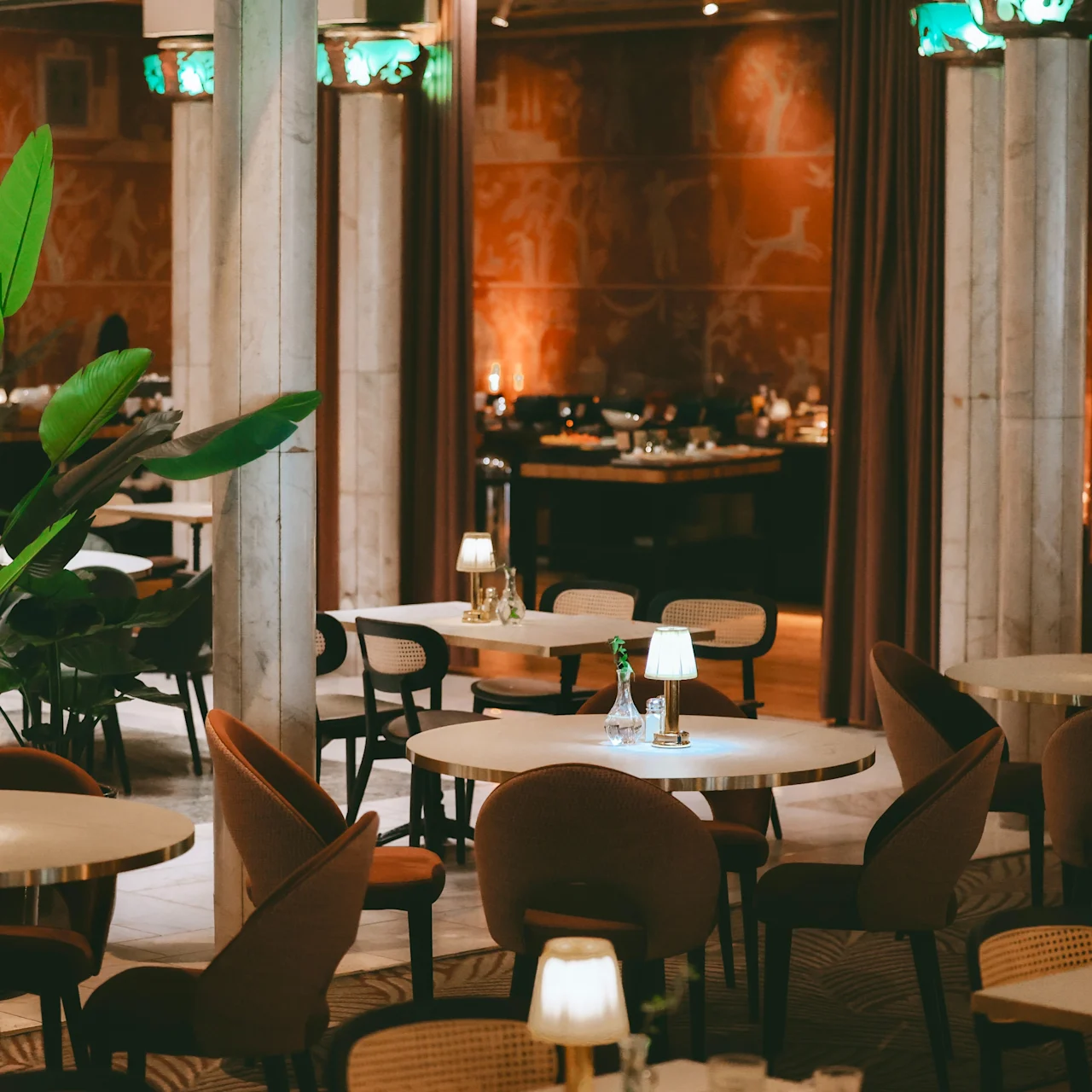 Tables are set with lamps and glasses in a warmly lit restaurant, surrounded by pillars and decorative plants, creating an inviting dining atmosphere. No text is present.