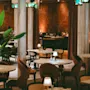 Tables are set with lamps and glasses in a warmly lit restaurant, surrounded by pillars and decorative plants, creating an inviting dining atmosphere. No text is present.