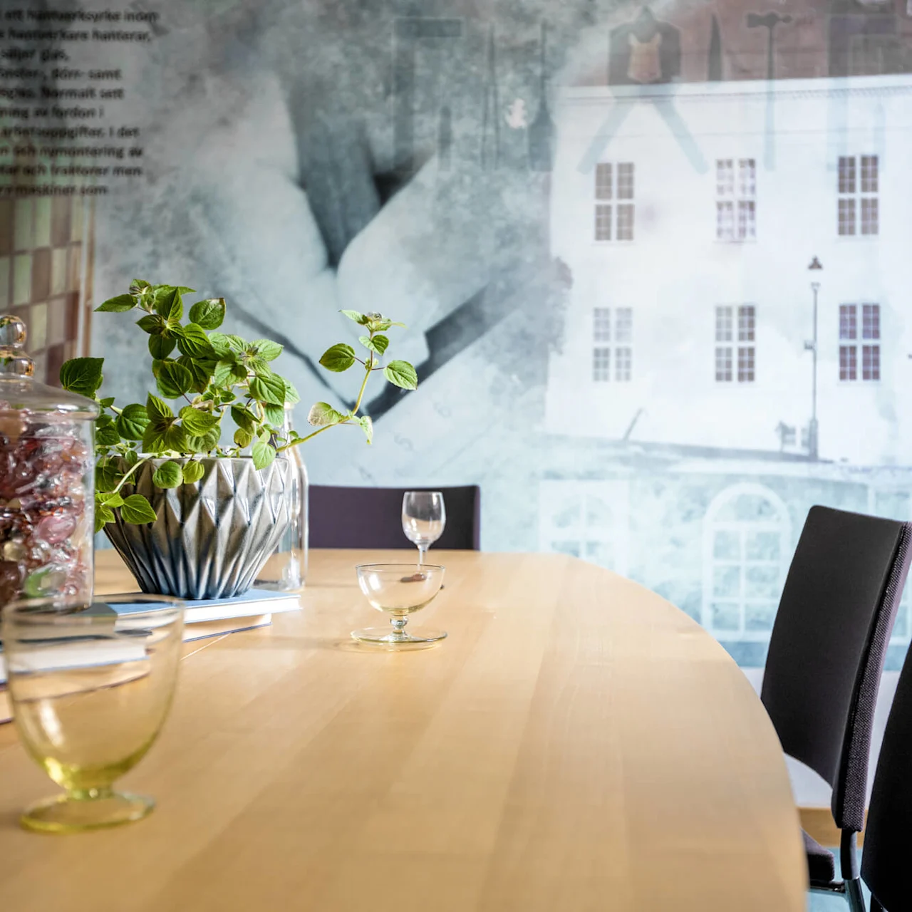 A wooden conference table with chairs, a potted plant, candy jar, books, and glasses, against a wall with a mural of a building.