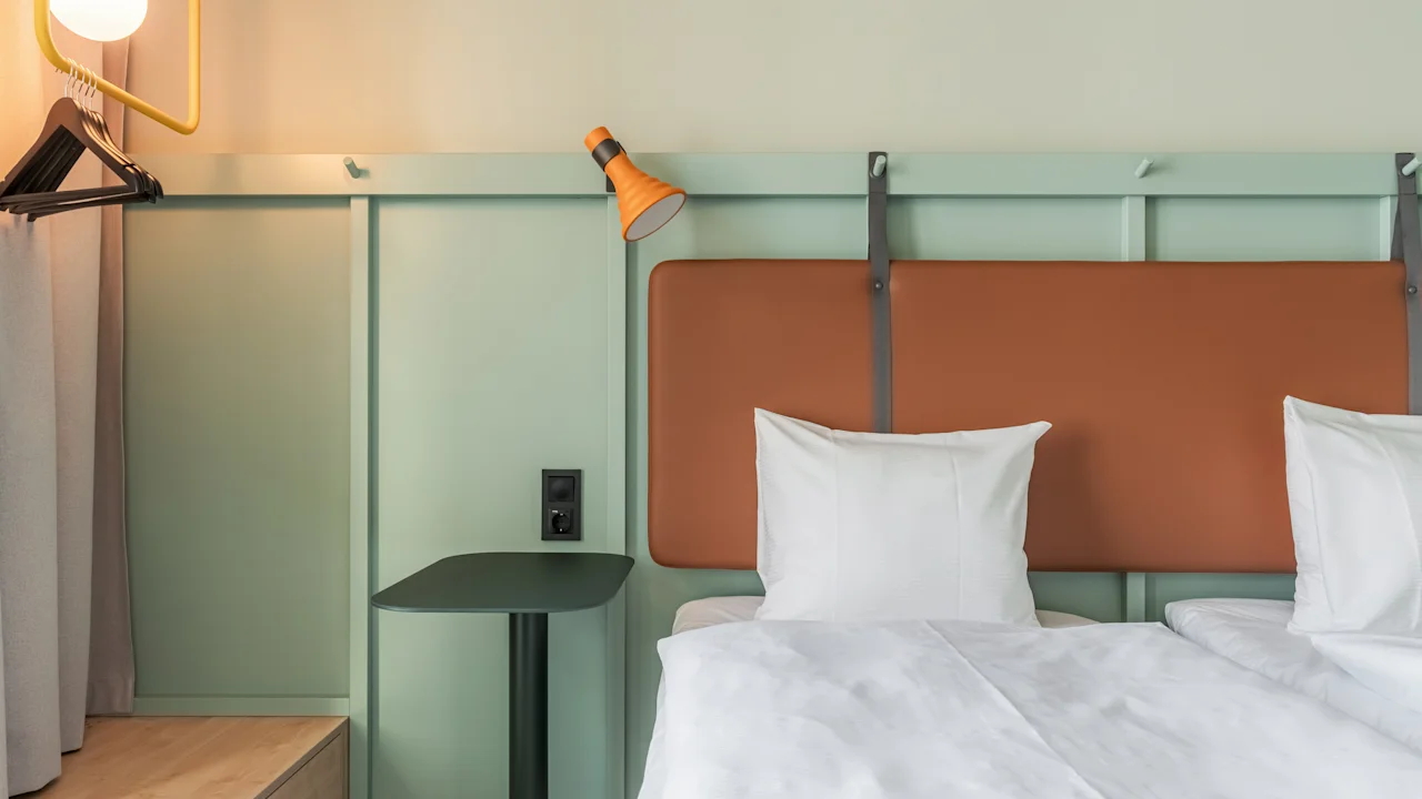 En pent oppredd seng med hvitt sengetøy og oransje sengegavl, akkompagnert av en oransje vegglampe. Et lite sidebord og hengende klær.