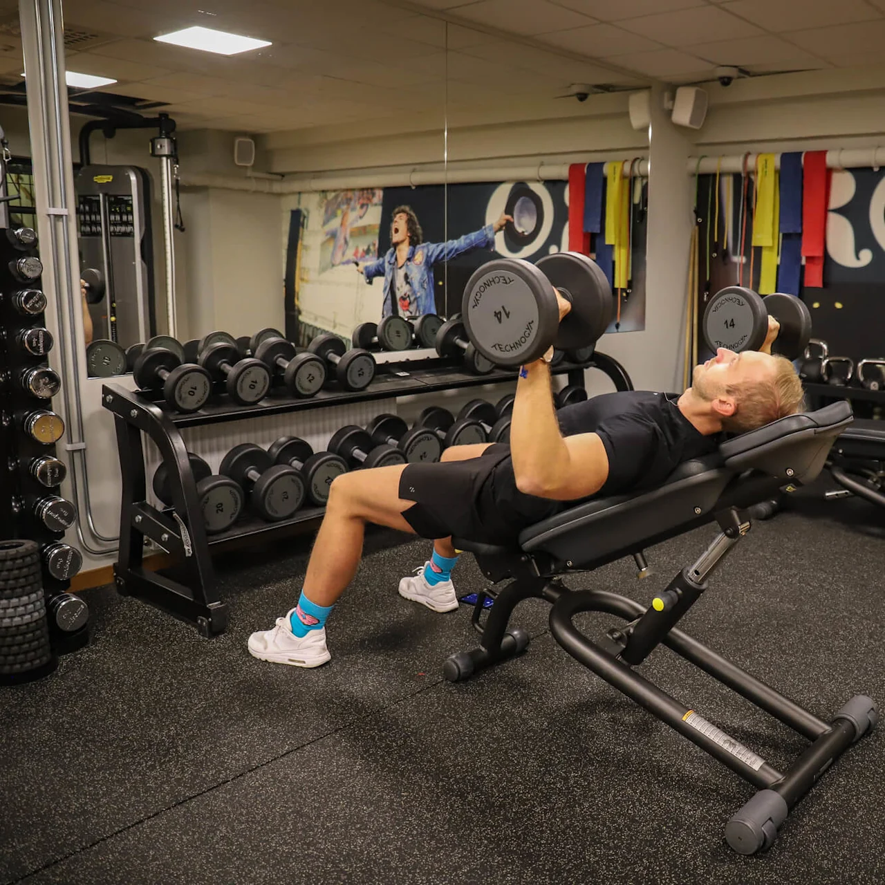 En man ligger på en lutande bänk och pressar hantlar i ett gym, omgivet av vikter och utrustning. En muralmålning av en person i en jeansjacka dekorerar väggen.