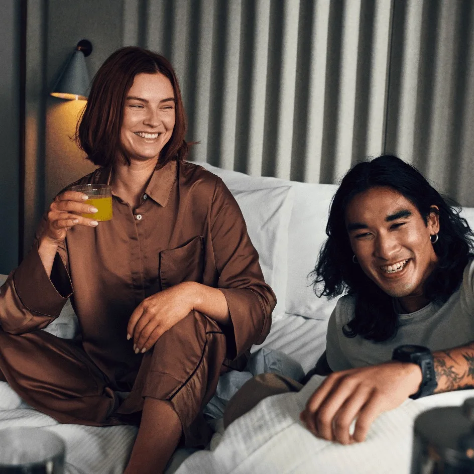 En person i silke-pyjamas holder et glas, smiler ved siden af en anden person med tatoveringer, liggende på en seng i et varmt oplyst rum med tøj synligt i et åbent garderobeskab.