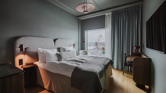 En seng med hvidt og gråt sengetøj står mod en væg, flankeret af natborde med lamper. Et vindue viser udsigt over en industrihavn med kraner.
