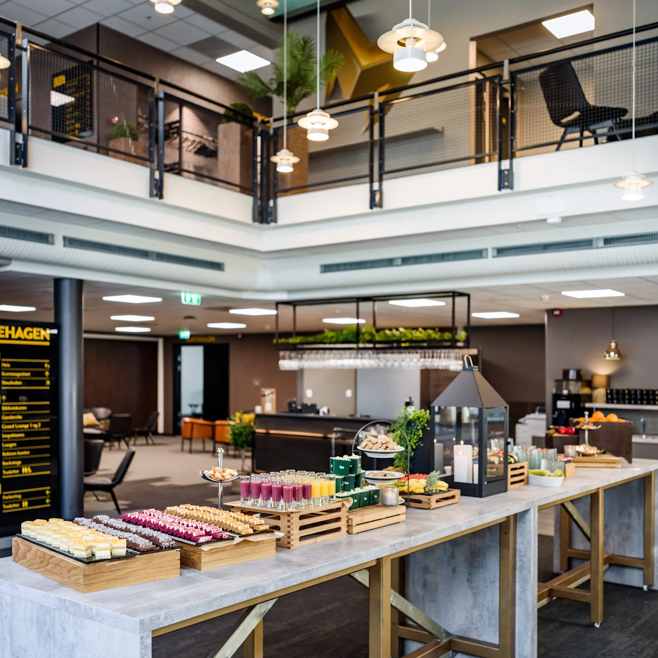 Buffet table displays assorted colorful snacks in a modern, open-concept dining area. Overhead lamps provide soft lighting, with stairs on the left leading to a mezzanine. Text: "EPLEHAGEN".
