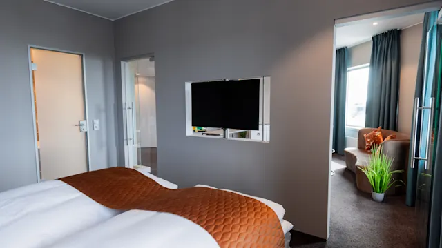 A neatly made bed with brown quilted accents sits in a modern hotel room, featuring wall-mounted TV and adjoining living area with a sofa and potted plant.