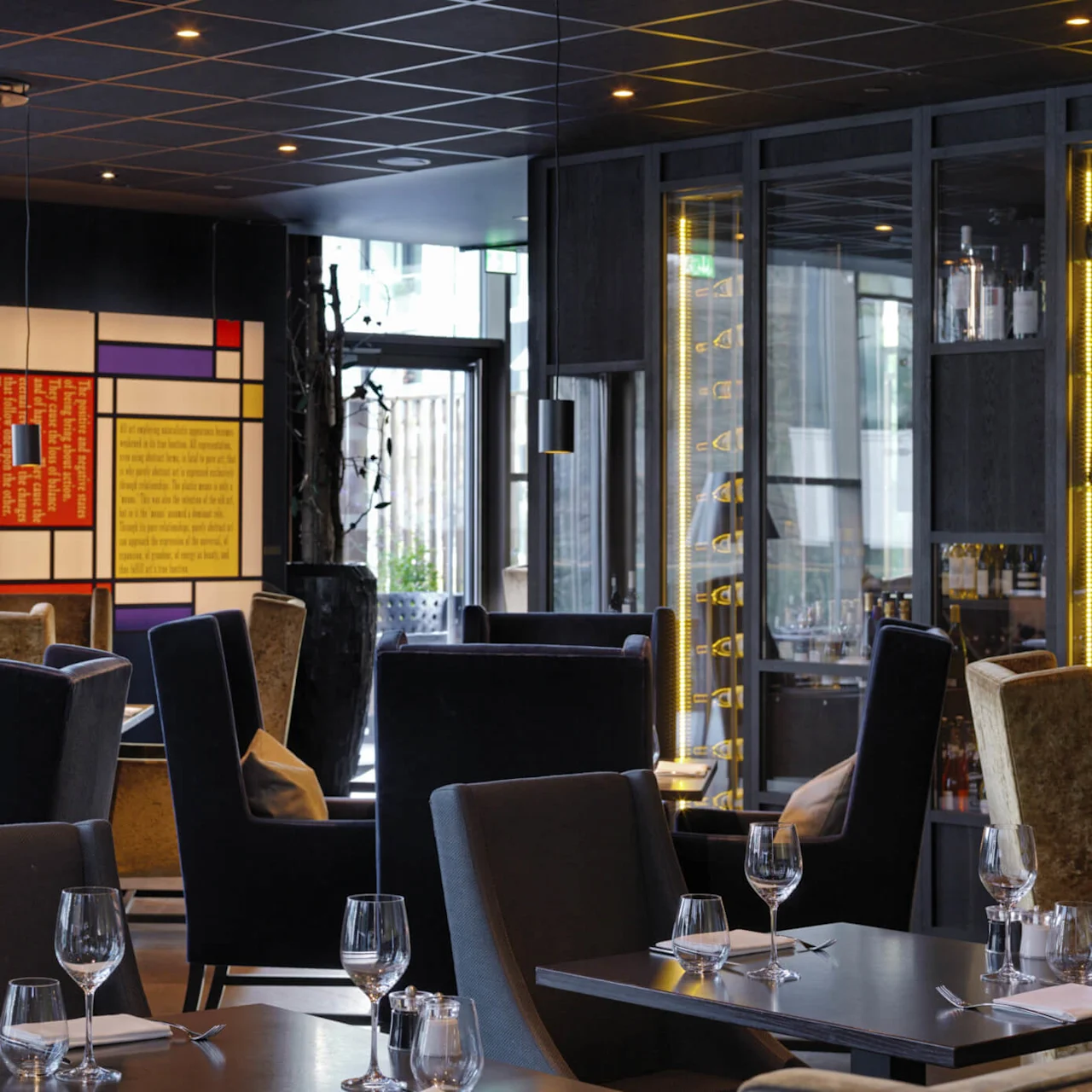 Restaurant med elegant, moderne innredning har pent dekkede bord med vinglass og servietter. Et fargerikt, abstrakt veggkunstverk tilføyer liv. Hyller viser flasker og forsterker den stilfulle atmosfæren.