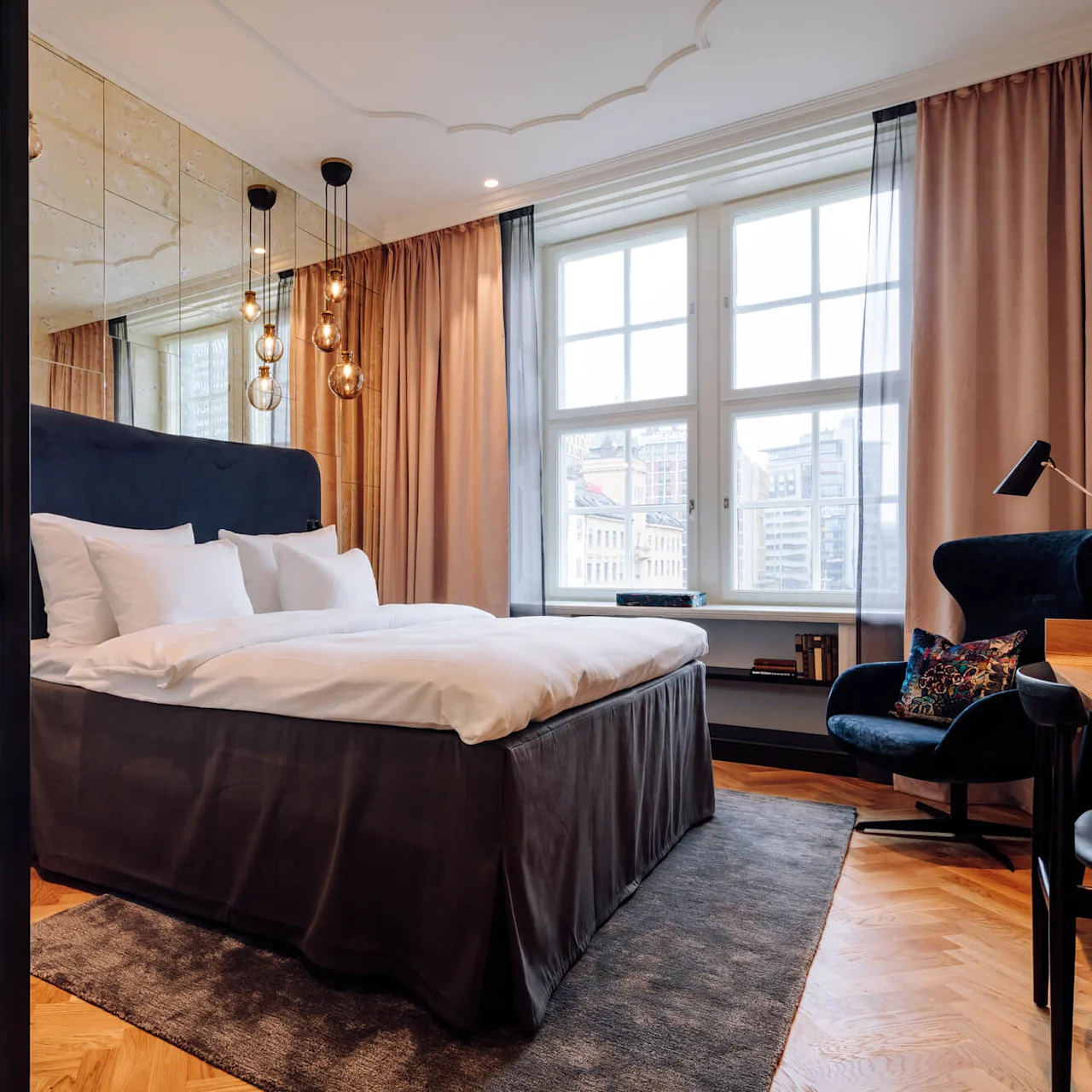 Bed with white linens, positioned against a mirrored wall, surrounded by hanging lights and large window, located in a modern room with wood flooring, chair, and desk.