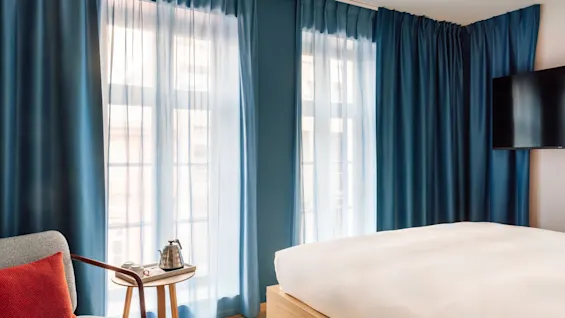 A comfortable hotel room with a bed, a cozy armchair, and a kettle for hot drinks. Blue curtains frame the window.