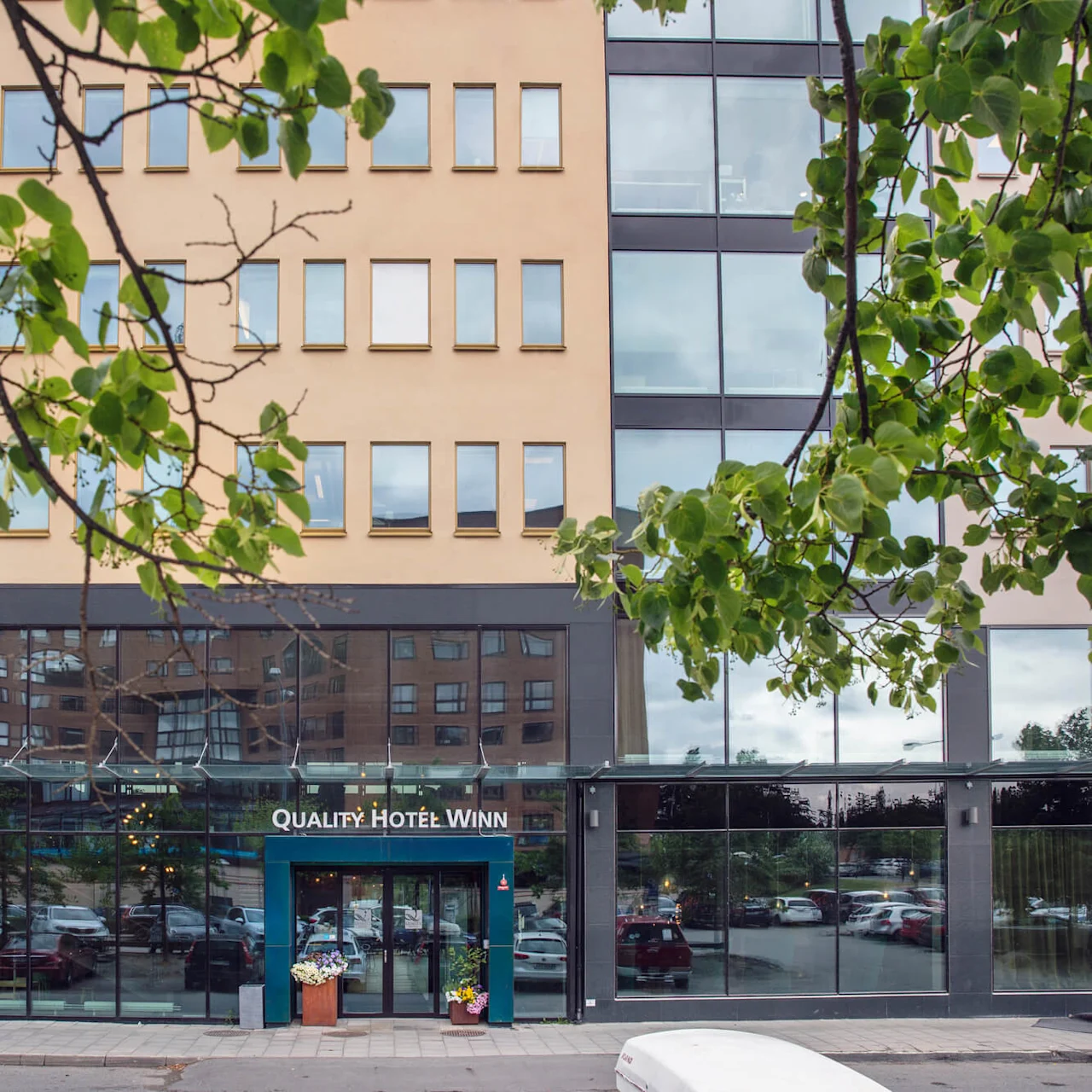 En hotellentré visar en skylt med texten "Quality Hotel Winn"; omgiven av stora fönster och grönska, med parkerade bilar synliga i reflektionen.
