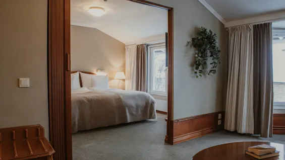 Ett mysigt hotellrum med ett sovrum synligt genom en öppen dörröppning, med en bekväm säng och ett ljust fönster.