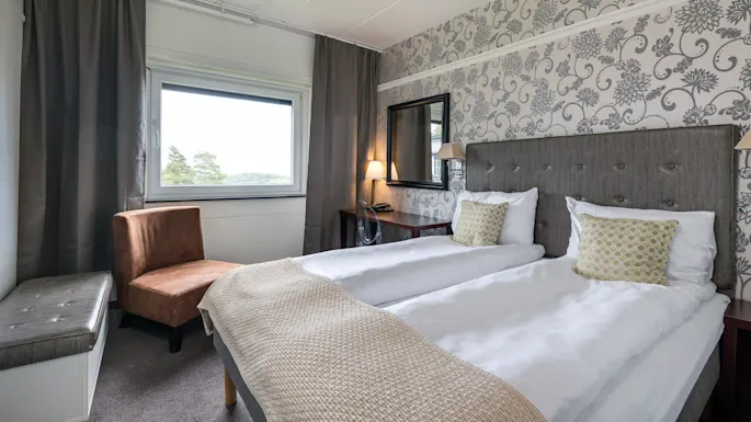 A neatly made twin bed with white linens and patterned cushions sits in a room with floral wallpaper, a window, a brown chair, and a nightstand with a lamp.