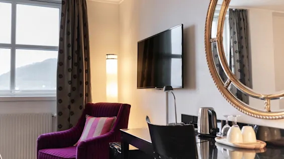Mukava hotellihuone, jossa on eloisa violetti nojatuoli, työpöytä virvokkeita varten ja suuri peili, joka heijastaa huoneen kutsuvaa tunnelmaa.