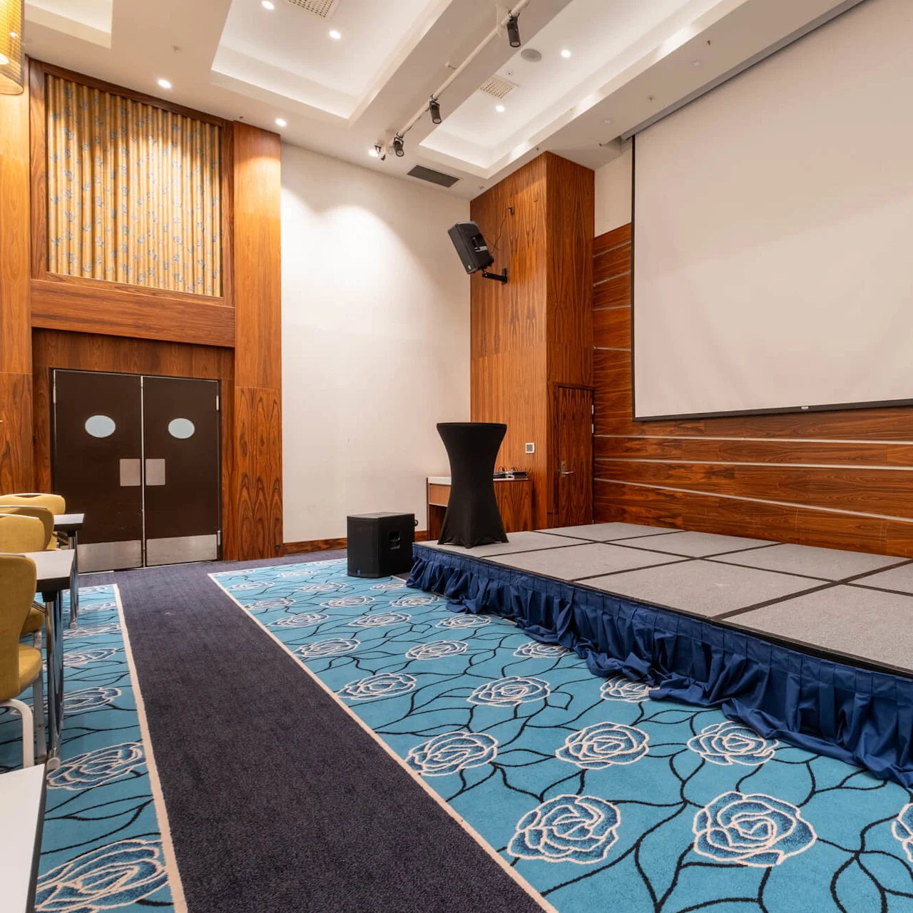 Stage with a black podium stands in a conference room featuring a large projector screen, wooden walls, and a blue patterned carpet. Speakers and chairs are positioned around.