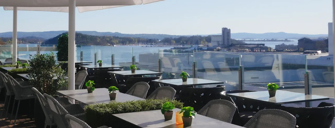 Outdoor terrace with tables and chairs overlooking a scenic waterfront with mountains and city buildings under a clear sky.