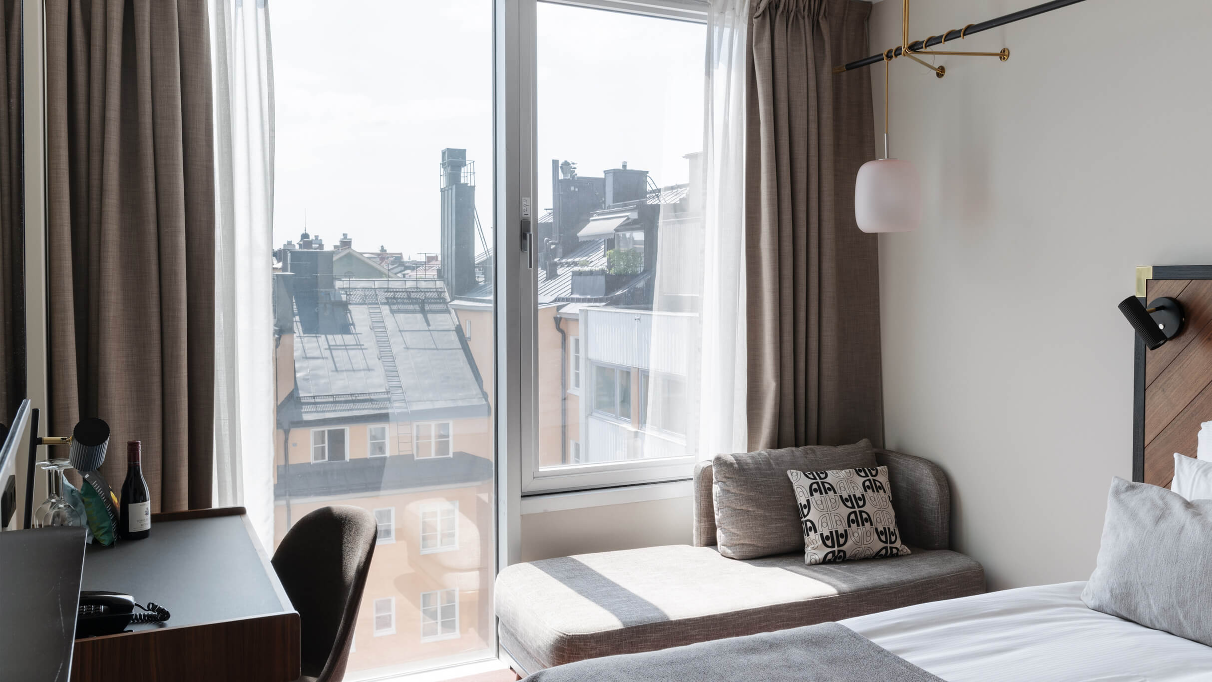 Nykyajan hotellihuone, jossa suuri ikkuna tarjoaa näkymät kaupunkimaisemaan, sisältäen sängyn, yöpöydän, kirjoituspöydän, tuolin ja minimalistisen sisustuksen.