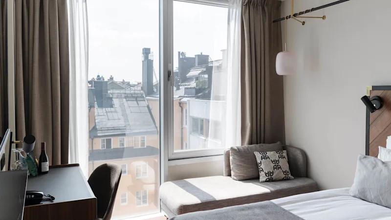 Et moderne hotelværelse med et stort vindue, der tilbyder udsigt over bylandskabet, har en seng, sengebord, skrivebord, stol og minimalistisk dekor.