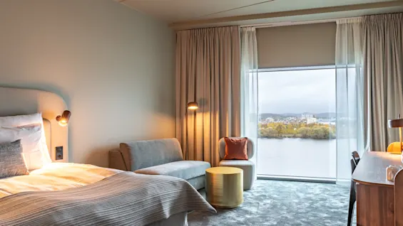 Ett mysigt hotellrum har en prydligt bäddad säng bredvid en liten soffa. Genom stora fönster syns en sjö och en avlägsen stadsbild.