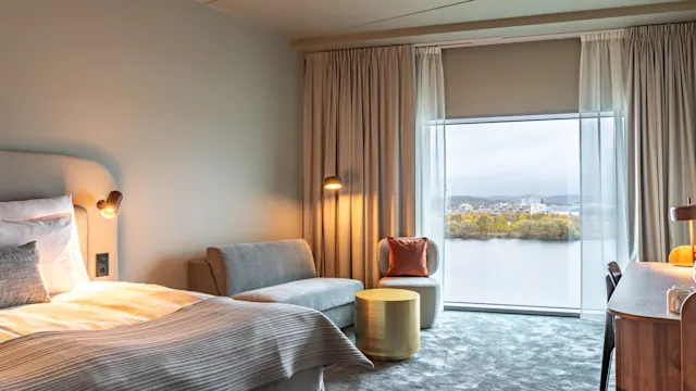 Et hyggeligt hotelværelse har en pænt redt seng ved siden af en lille sofa. Gennem store vinduer ses en sø og en fjern bysiluet.