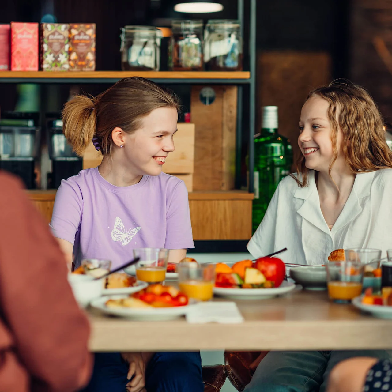 Two children sit smiling at a dining table with plates of food and glasses of orange juice, inside a cozy room with shelves of jars and boxes in the background. 